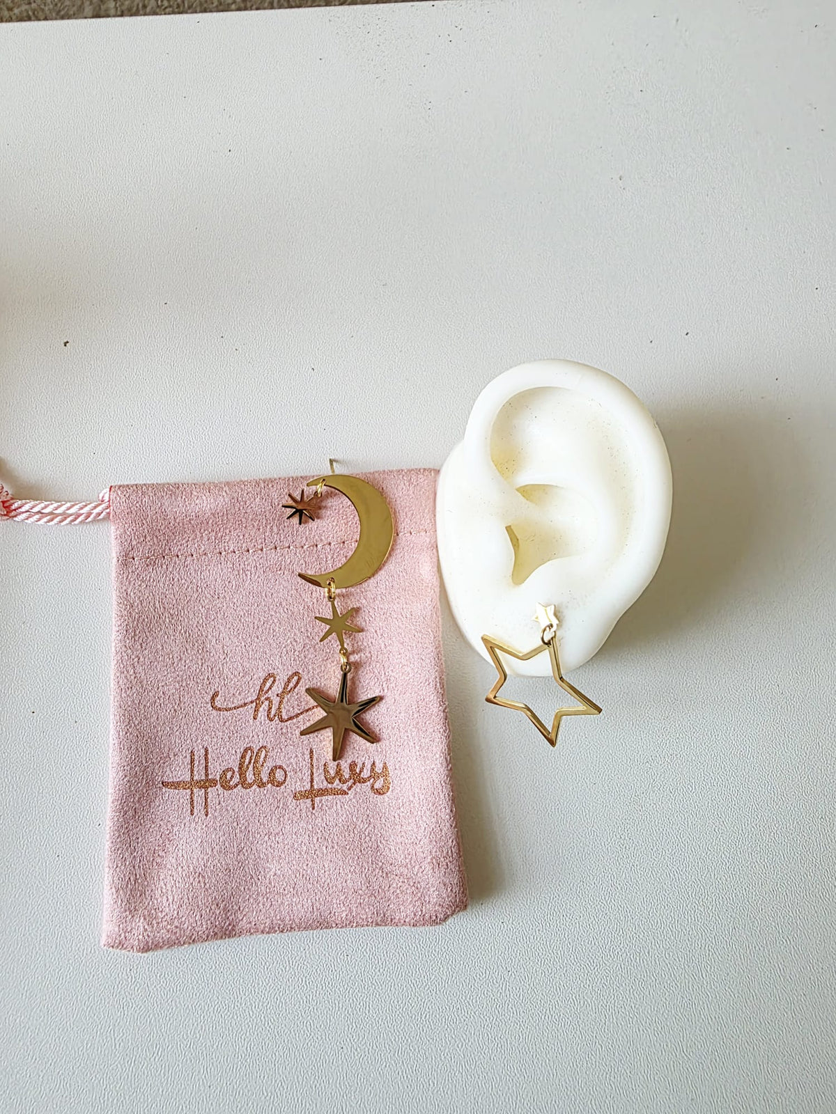 Gold star and moon earring on a white ear model with a pink pouch labeled 'Hello Luxy' on a light gray background.