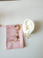 Gold star and moon earring on a white ear model with a pink pouch labeled 'Hello Luxy' on a light gray background.