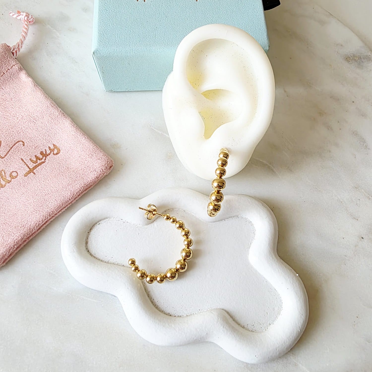 Gold hoop earrings on a white ear model with 'Hello Luxy' box and pouch in the background.