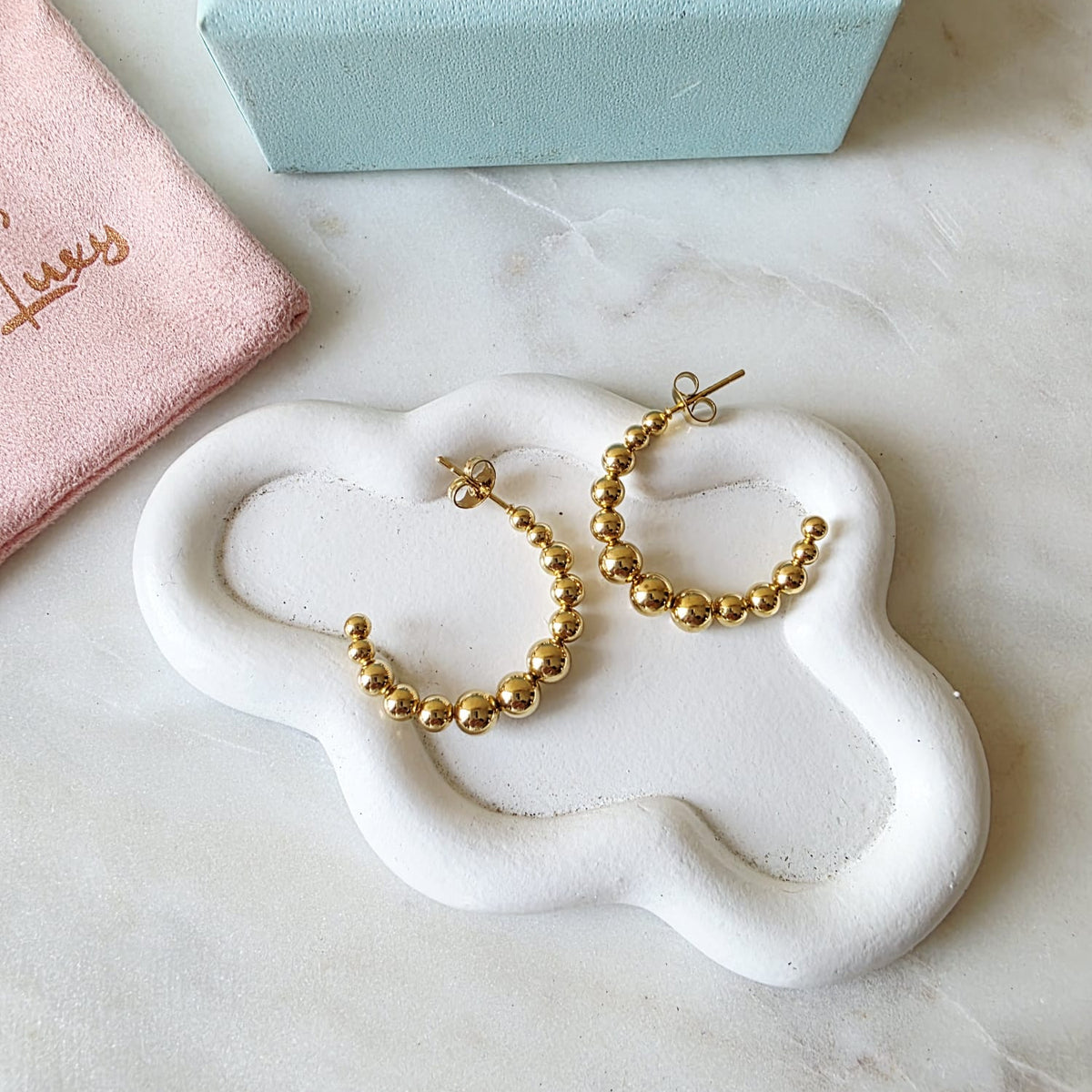 Gold hoop earrings on a white cloud-shaped stand with a pink jewelry box in the background.