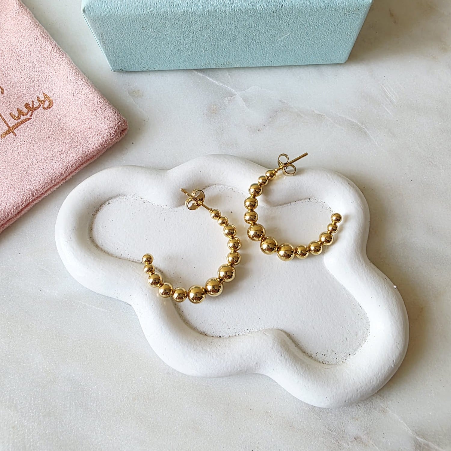 Gold hoop earrings on a white cloud-shaped stand with a pink jewelry box in the background.