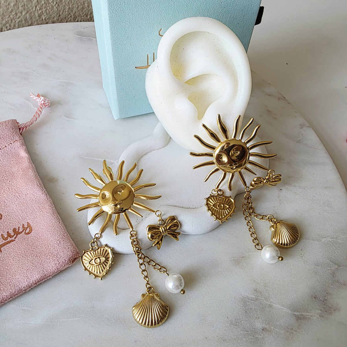 Gold sun-shaped earrings with pearl accents on a white ear model, placed on a marble surface.